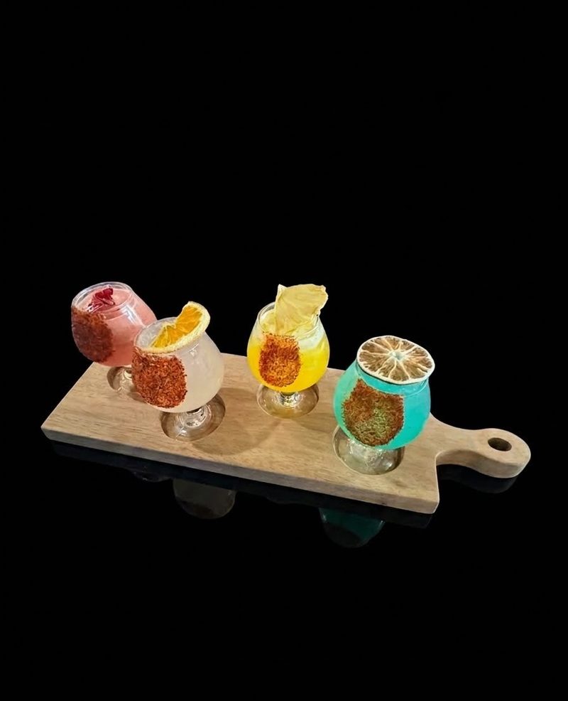 A wooden board with a flight of four colorful drinks with chile-rimmed glasses